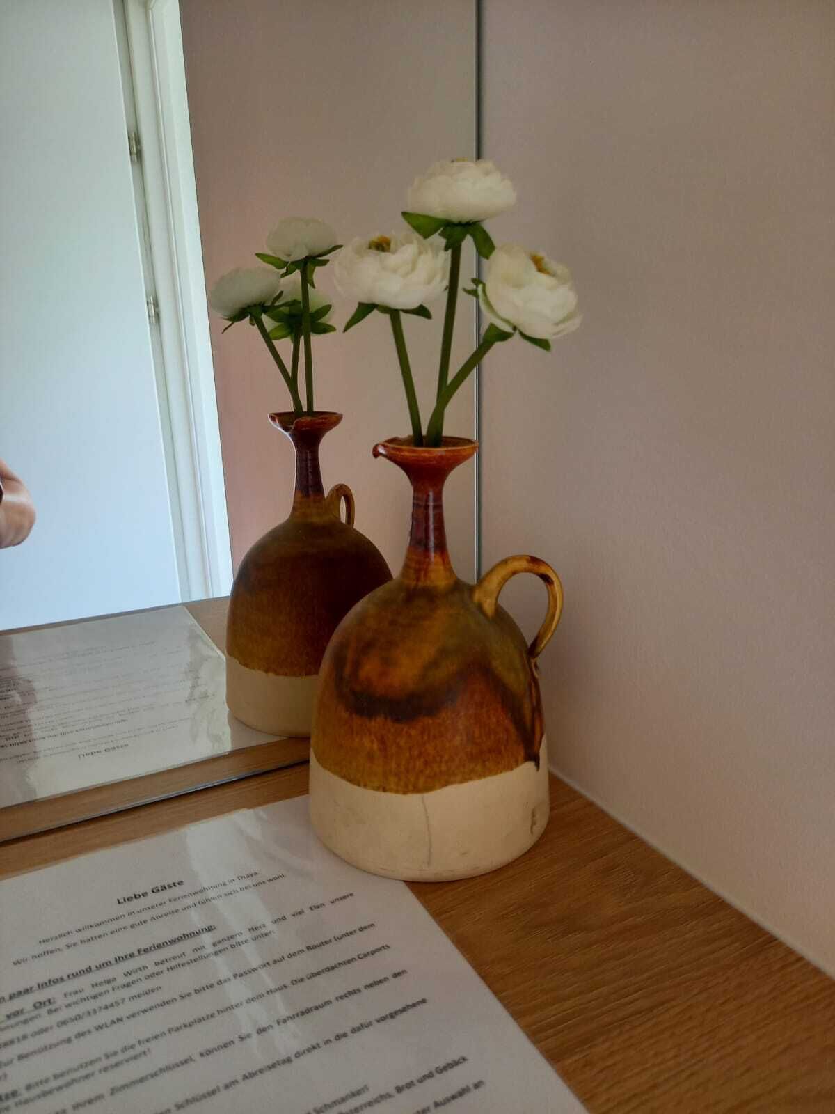Dos jarrones de cerámica con flores blancas sobre una mesa de madera, un espejo que refleja los jarrones y un menú delante del espejo.
