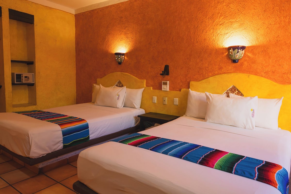 A hotel room with two beds, each with white bedding and colorful blankets, a wooden headboard, and wall-mounted light fixtures.