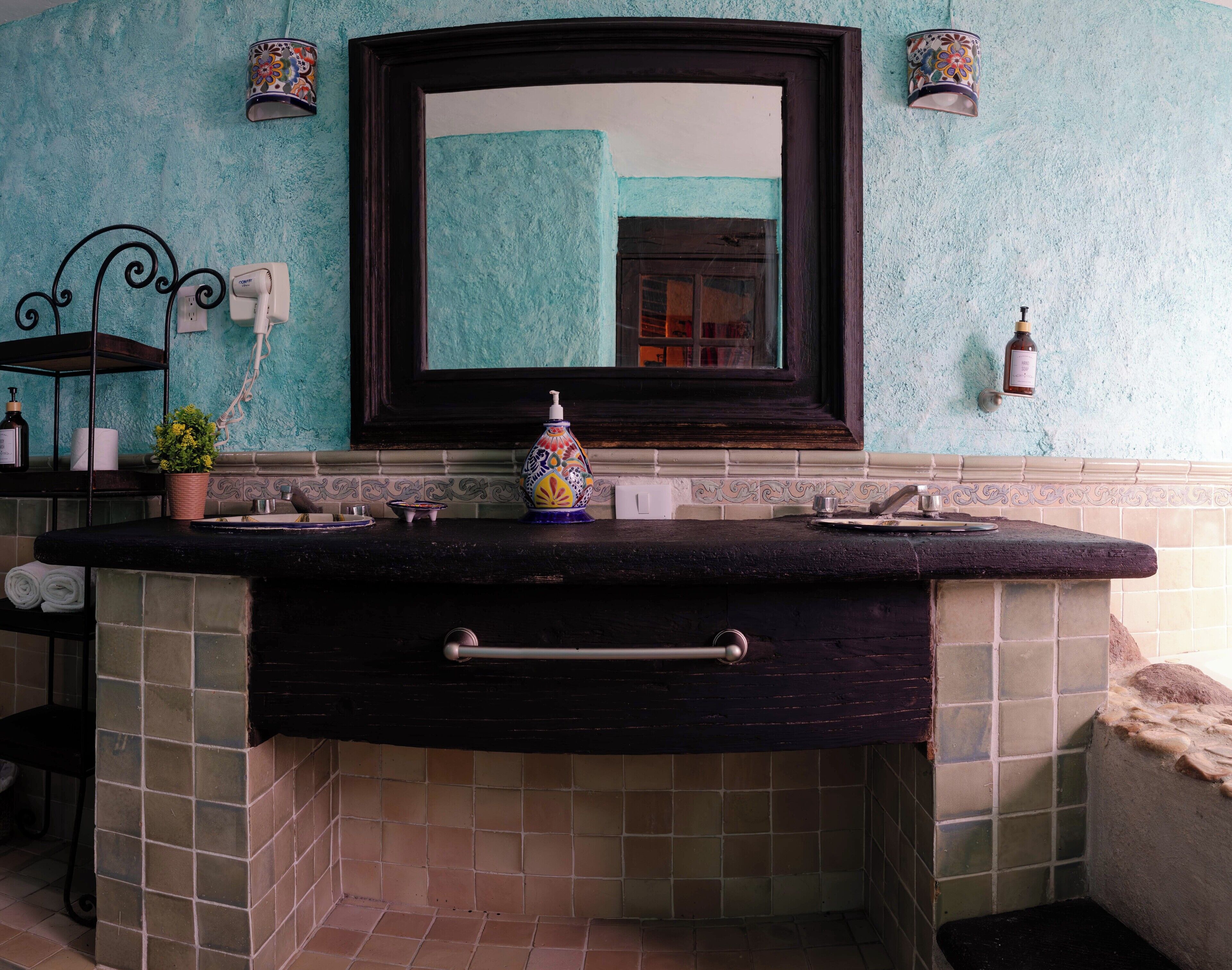 Un baño con encimera negra, un espejo y un lavabo. Hay dos luces empotradas en la pared y una botella de jabón para manos.