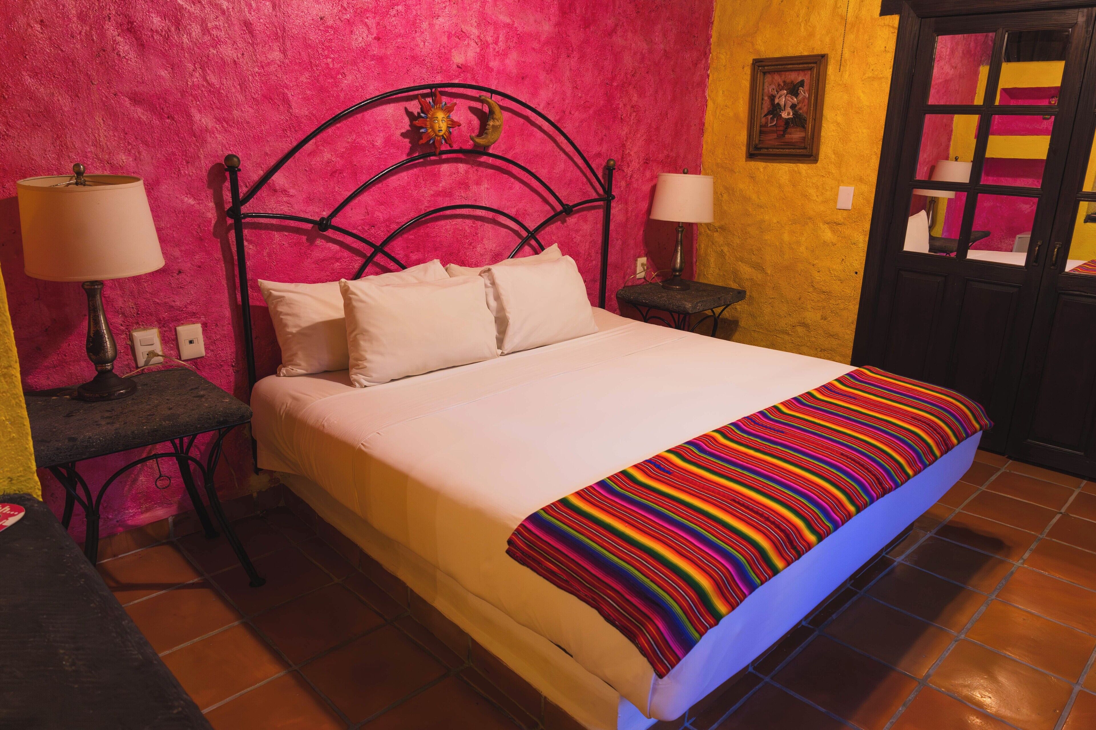 A bed with a white mattress and pillows, a colorful striped blanket, and a black metal headboard. There are two bedside tables with lamps. The walls are painted pink and yellow, and there is a door with a glass panel.