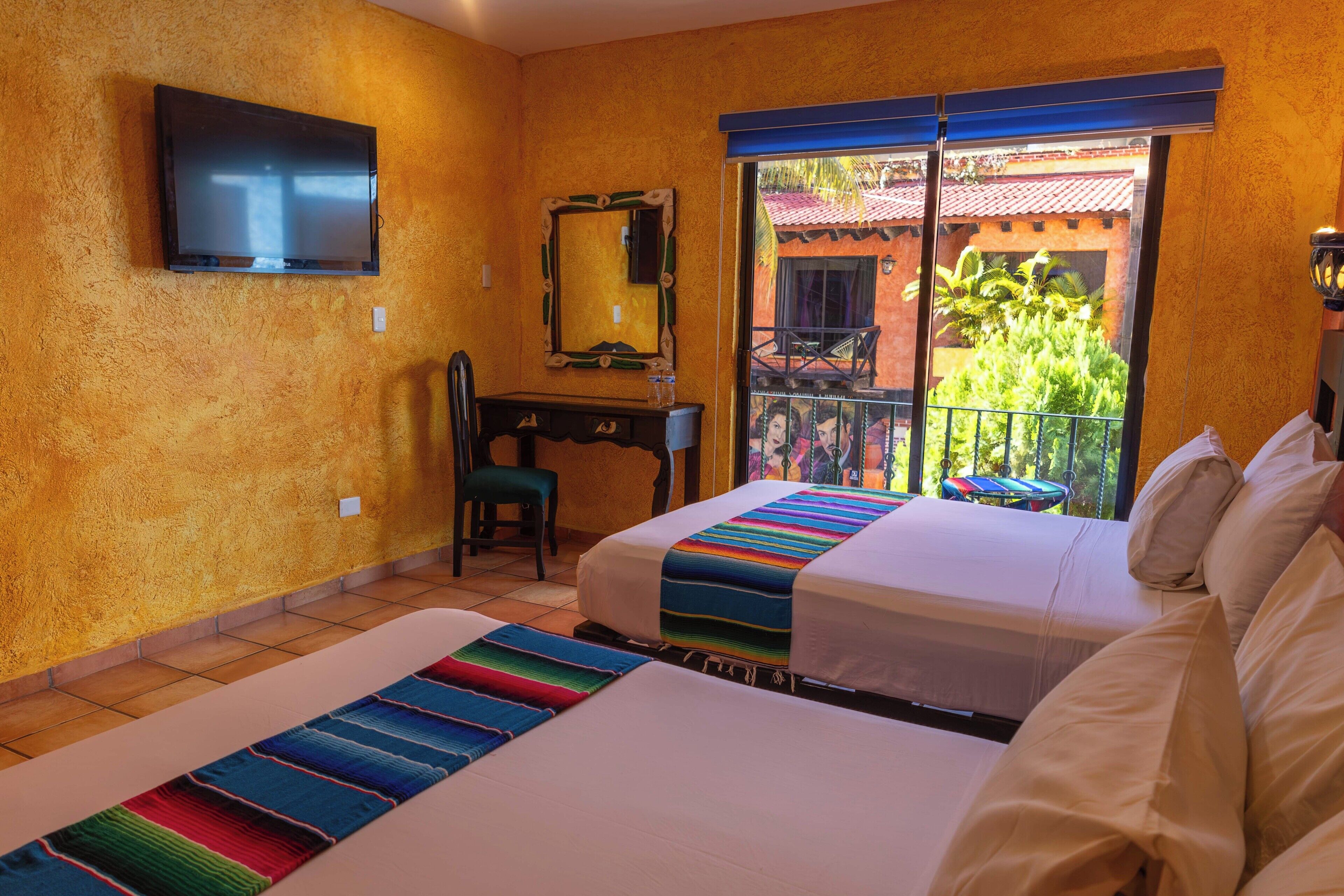 Habitación de hotel con dos camas, un escritorio, una silla, televisión y balcón con vistas a zonas verdes y un edificio.
