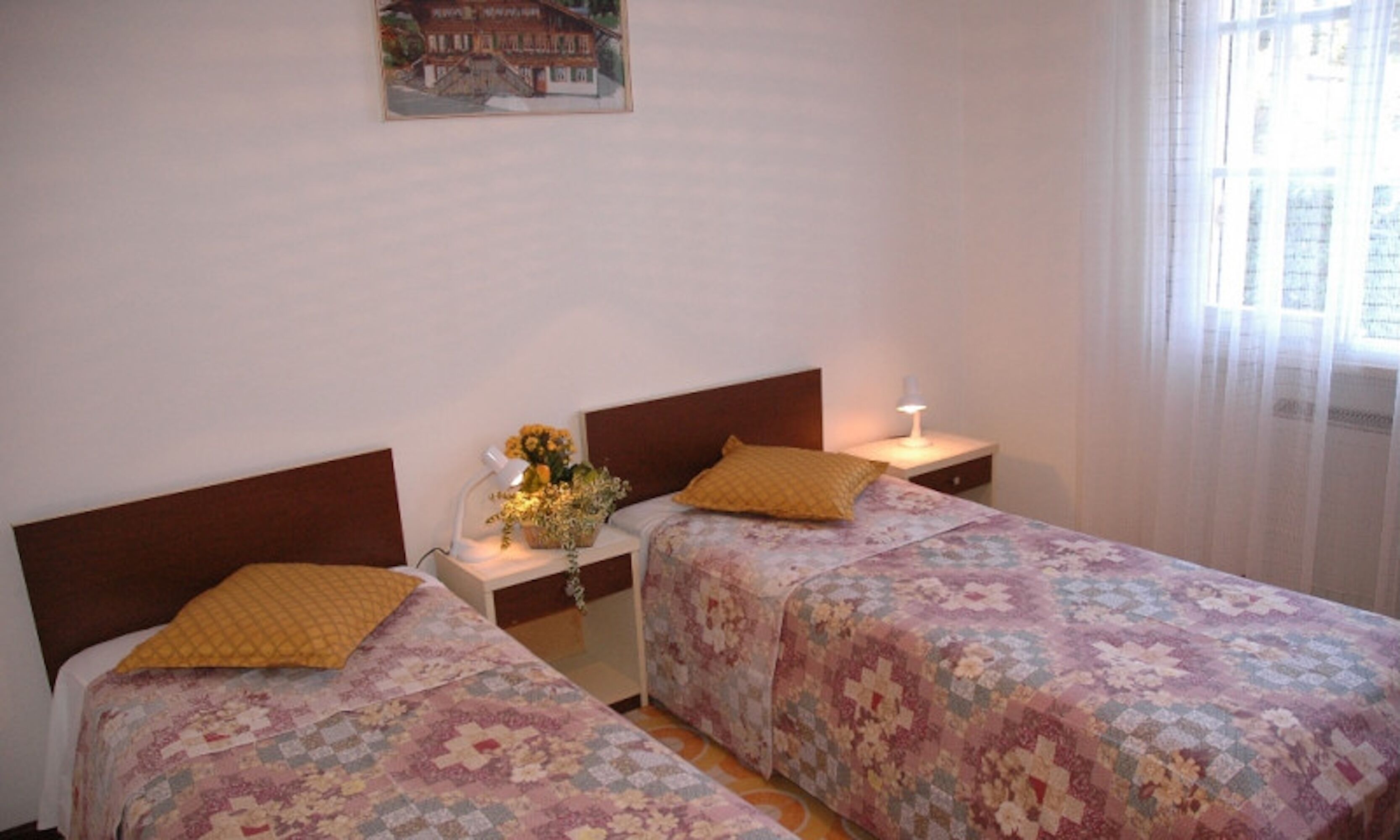 Una stanza con due letti, ciascuno con una testiera in legno e una coperta a fiori. Tra i letti, un comodino con una lampada e un vaso di fiori. Sulla parete sopra i letti è appeso un poster.