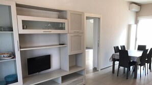 A modern living room with a flat-screen TV, built-in shelves, and a dining area with a table and chairs. - Sicily house 100 mt from the sea, garden, climate, terrace, laundry room (Cervia)