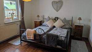 Ein Schlafzimmer mit einem Metallbettgestell, zwei Nachttischen mit Lampen, einem fenster mit Vorhängen, einer Heizung und einer herzförmigen Dekoration an der Wand.