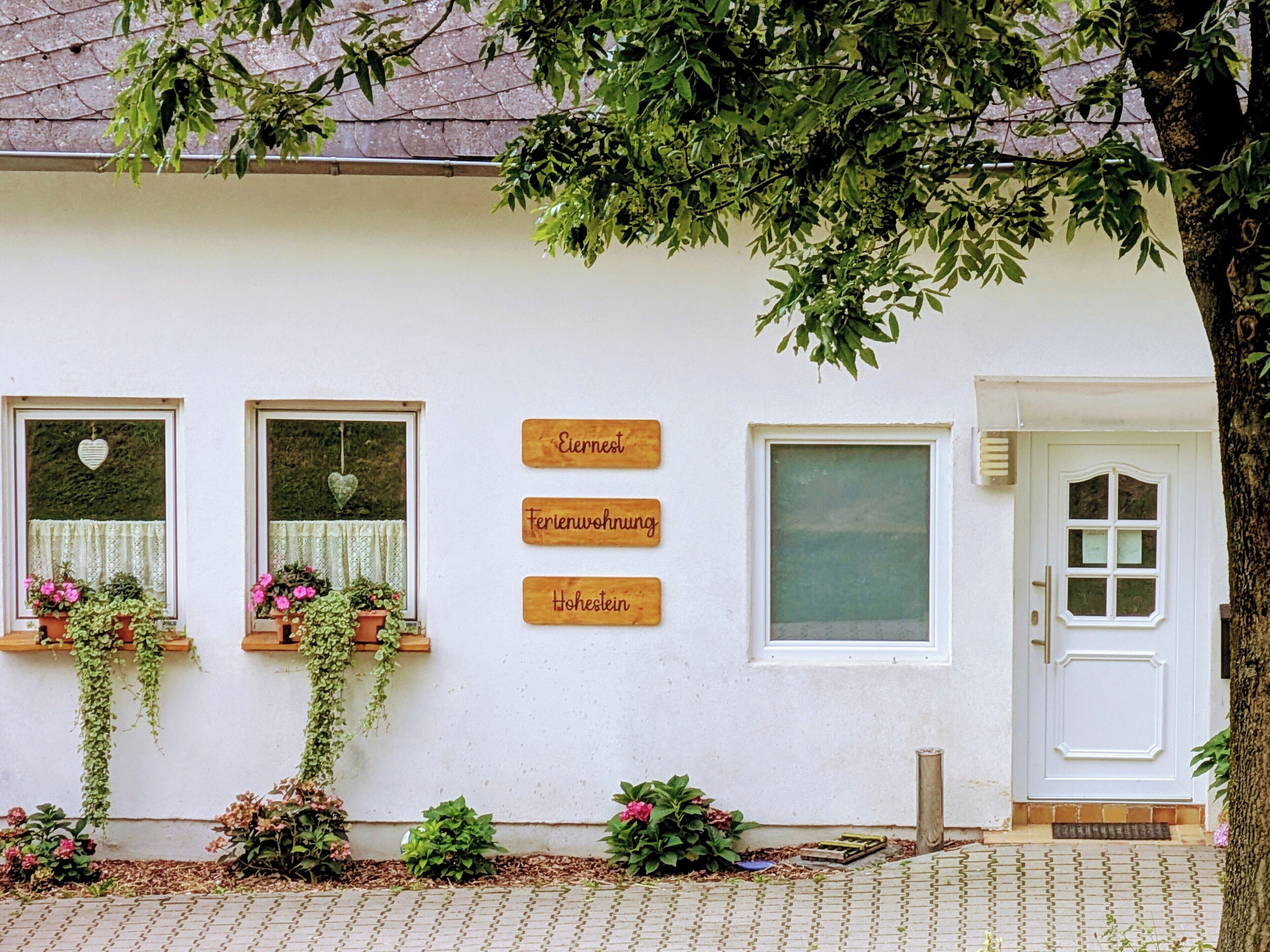 A white building with three windows, each with a heart-shaped decoration and hanging plants. There are three wooden signs on the wall reading "Eiernest," "Ferienwohnung," and "Hohestein."