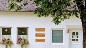 Ein weißes Gebäude mit drei Fenstern, jedes mit einer herzförmigen Dekoration und hängenden Pflanzen. Drei Holzschilder an der Wand zeigen die Aufschriften 'Eiernest', 'Ferienwohnung' und 'Hohestein'.