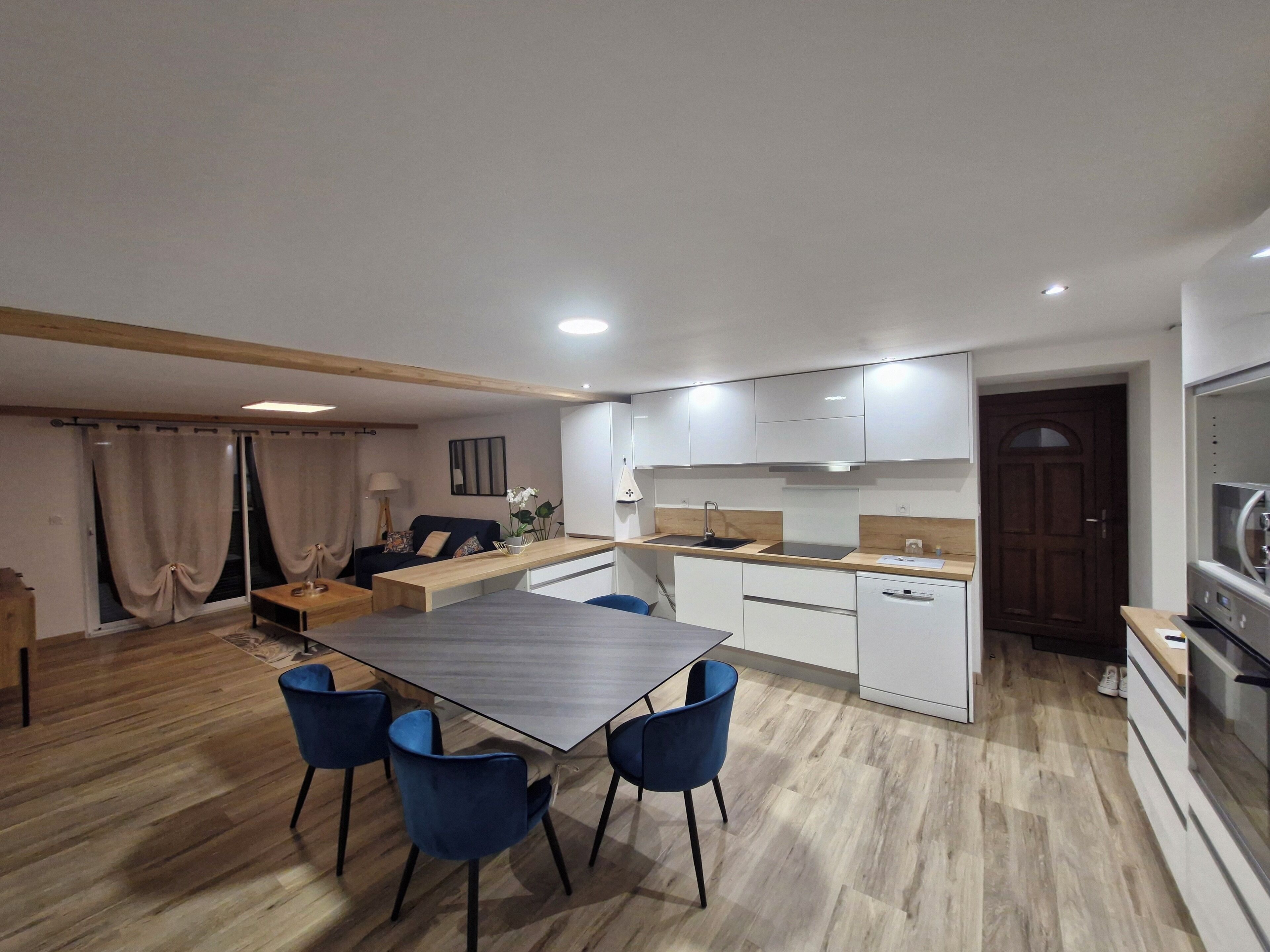 Cozinha moderna com armários brancos, bancada de madeira e forno embutido. Há uma área de refeições com uma mesa e cadeiras azuis.