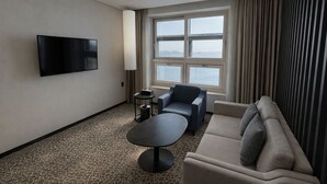 Een hotelkamer met een flatscreen televisie aan de muur, een groot raam met gordijnen, een bank, een stoel en een klein rond tafeltje.