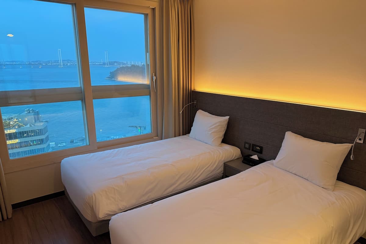 A hotel room with two single beds, a nightstand with a phone and a lamp, and a large window offering a view of a cityscape and water.