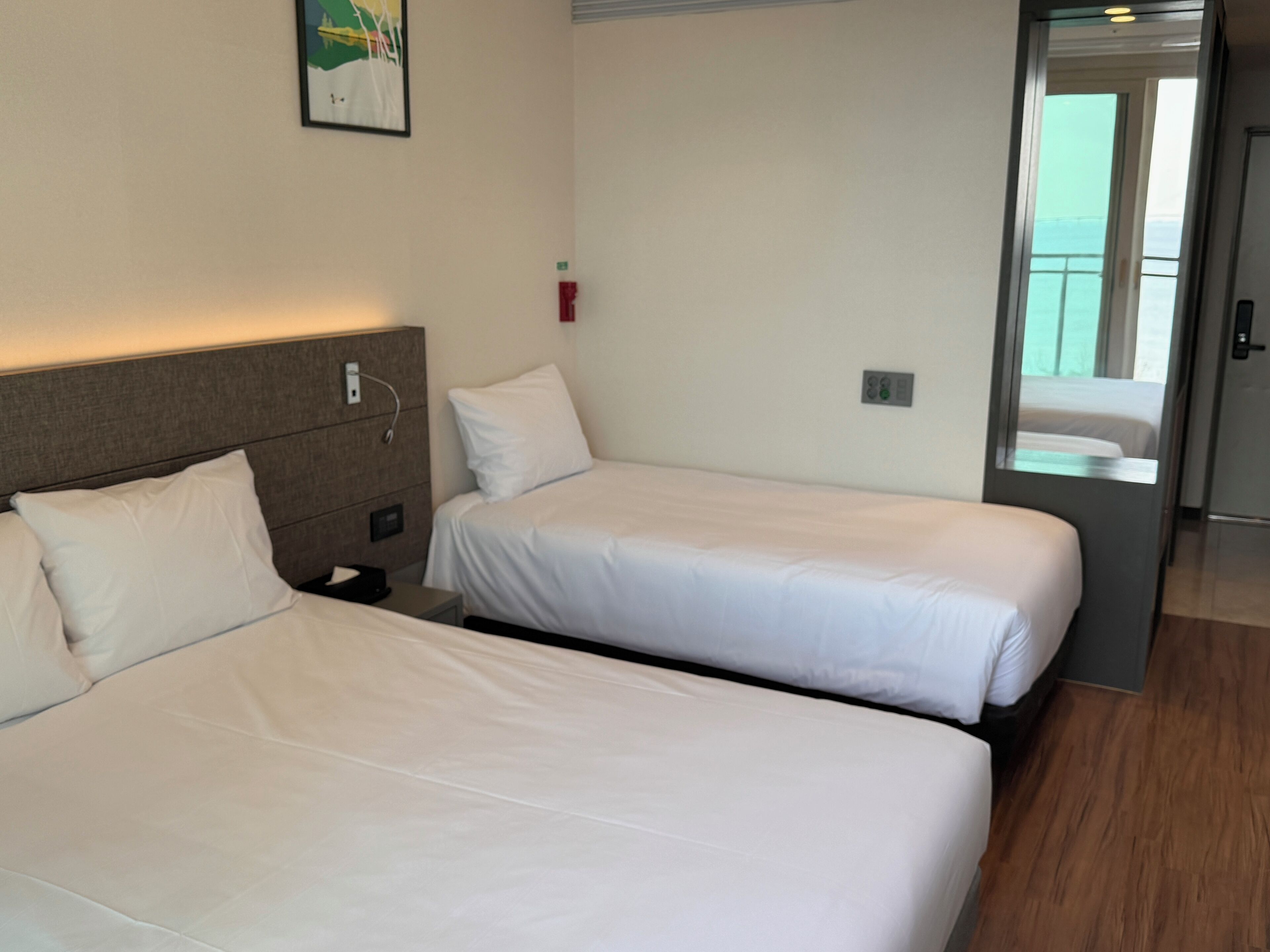 Une chambre d’hôtel avec deux lits simples, une tête de lit en bois, une petite table de chevet et une lampe fixée au mur.