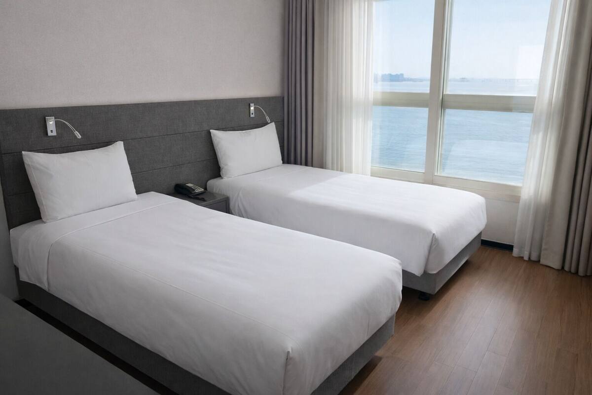 A hotel room with two beds, a bedside table with a phone, and a view of the sea through large windows.