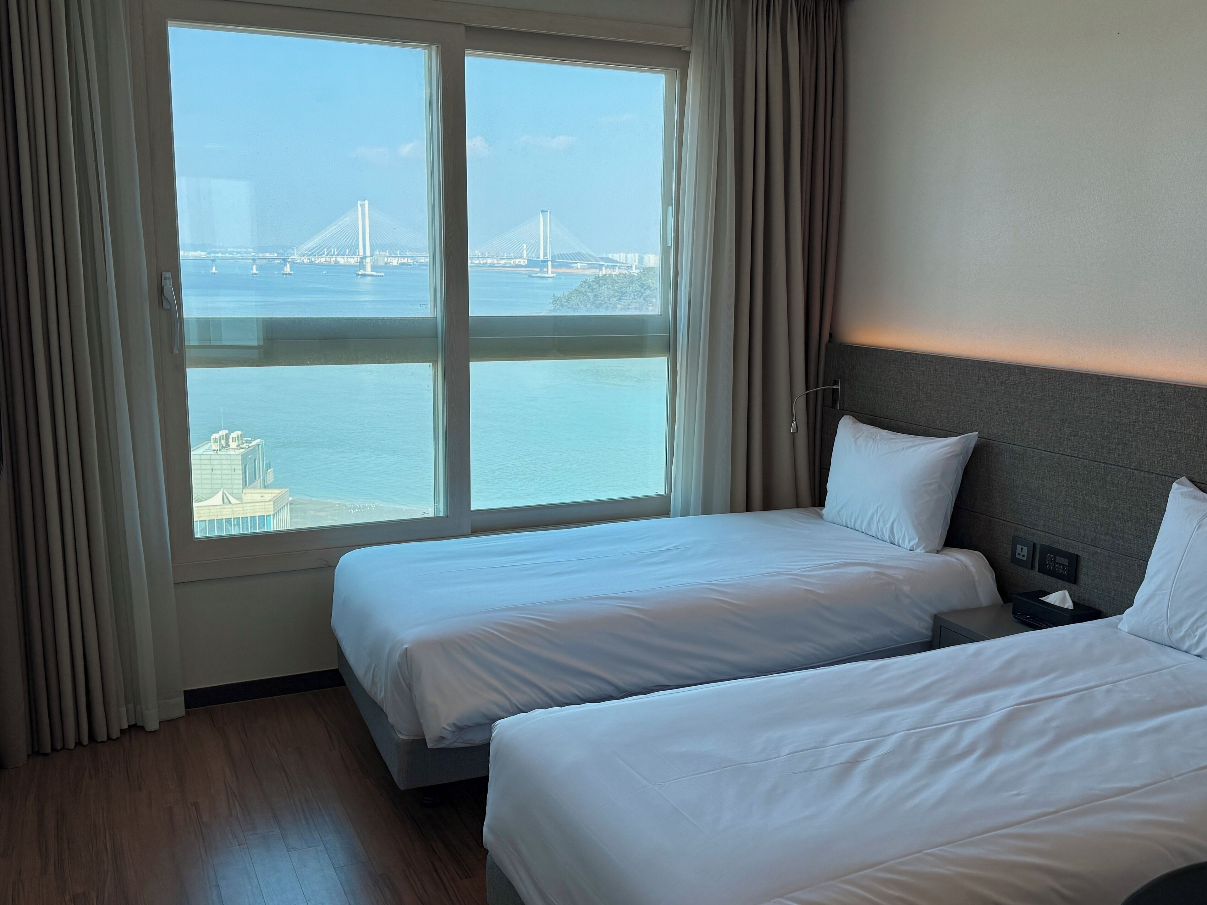 A hotel room with two beds, a large window with a view of a bridge and water, and a nightstand with a lamp.