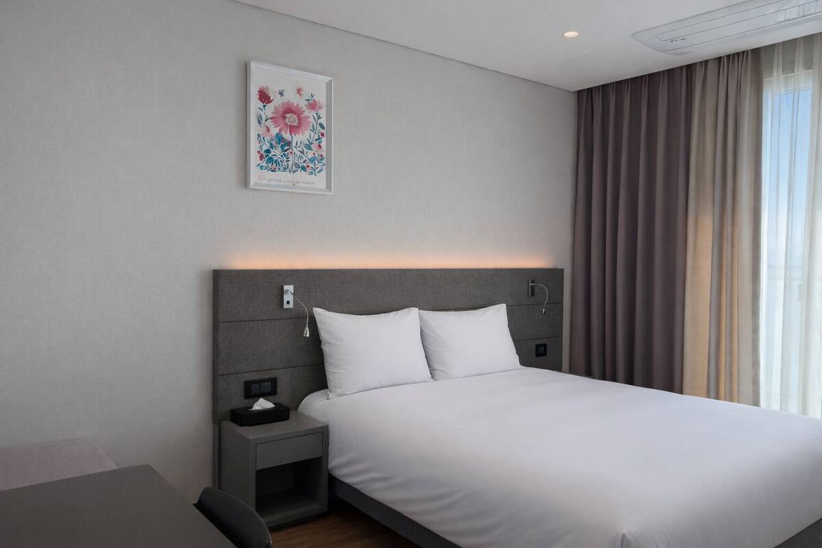 A modern hotel room with a large bed, a bedside table with a lamp, a small desk, and a window with curtains.