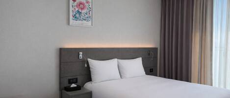 A modern hotel room with a large bed, a bedside table with a lamp, a small desk, and a window with curtains.