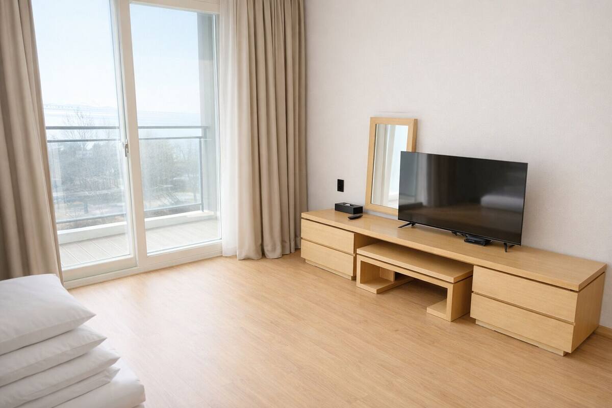 A modern hotel room with a wooden desk, a flat-screen TV, a large mirror, and a sliding glass door to the balcony.