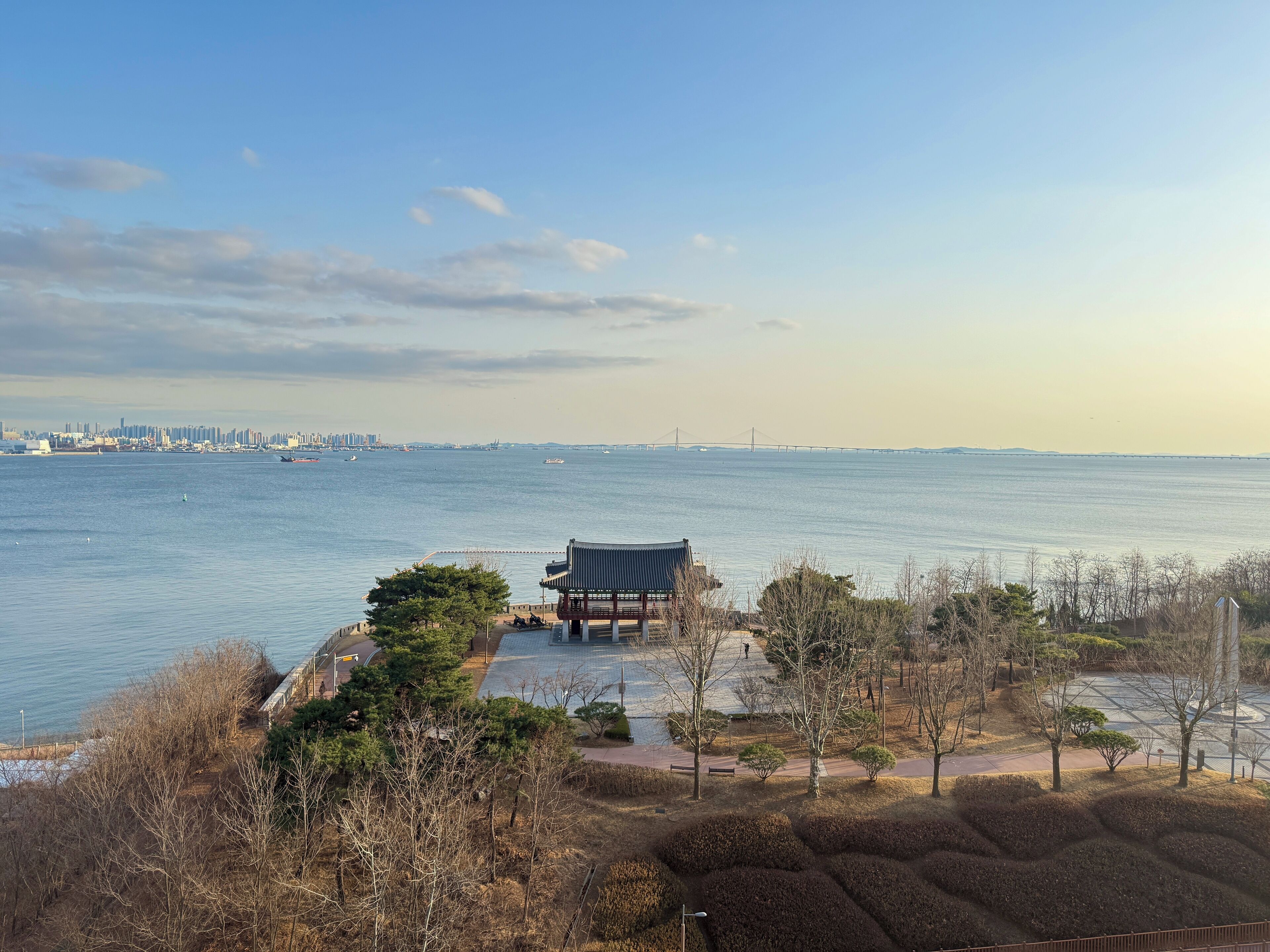 A riverside park with a traditional pavilion, pathways, and a distant city skyline.