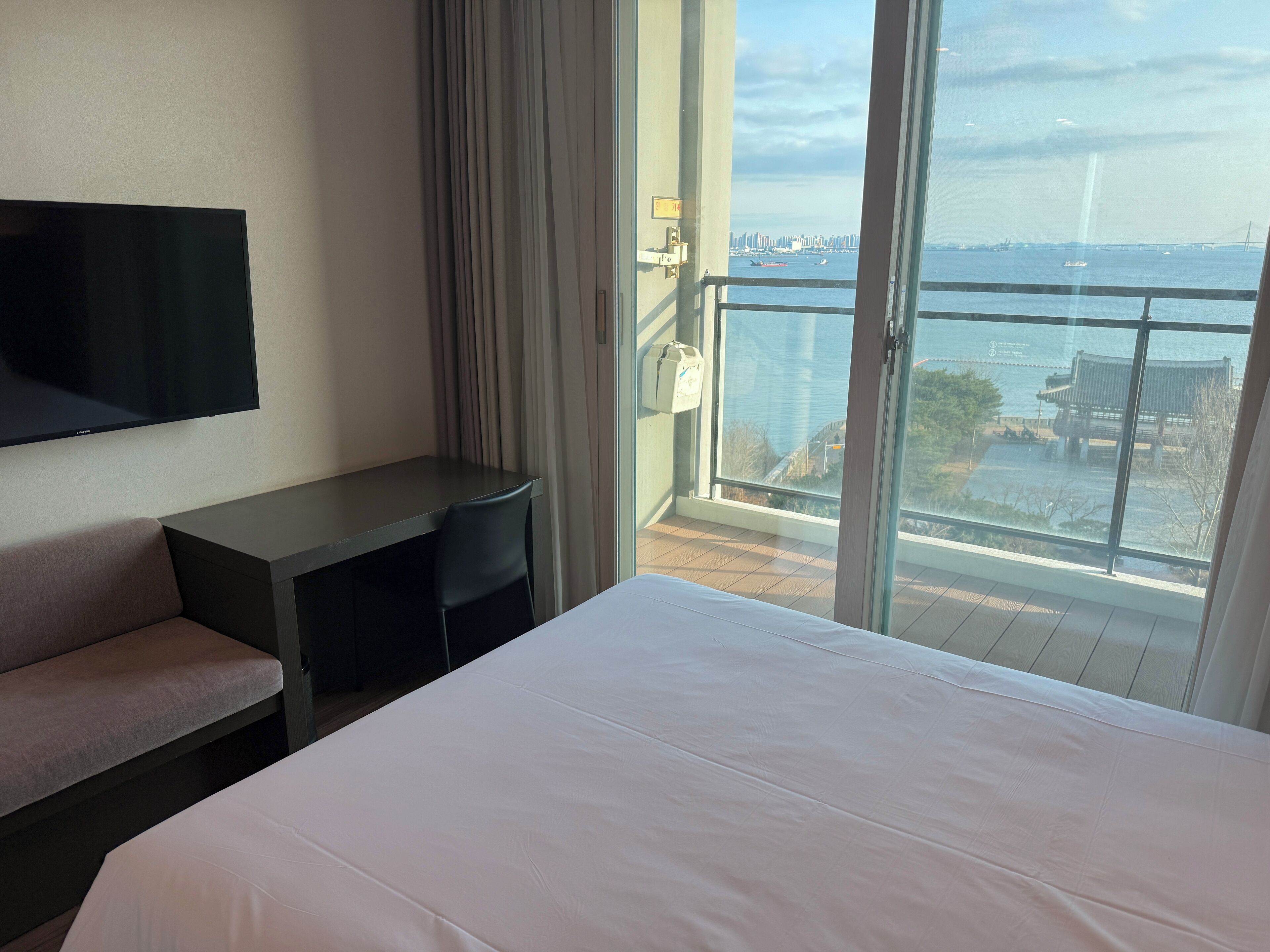 A hotel room with a bed, a desk, a chair, a TV, and a balcony with a view of the city and water.