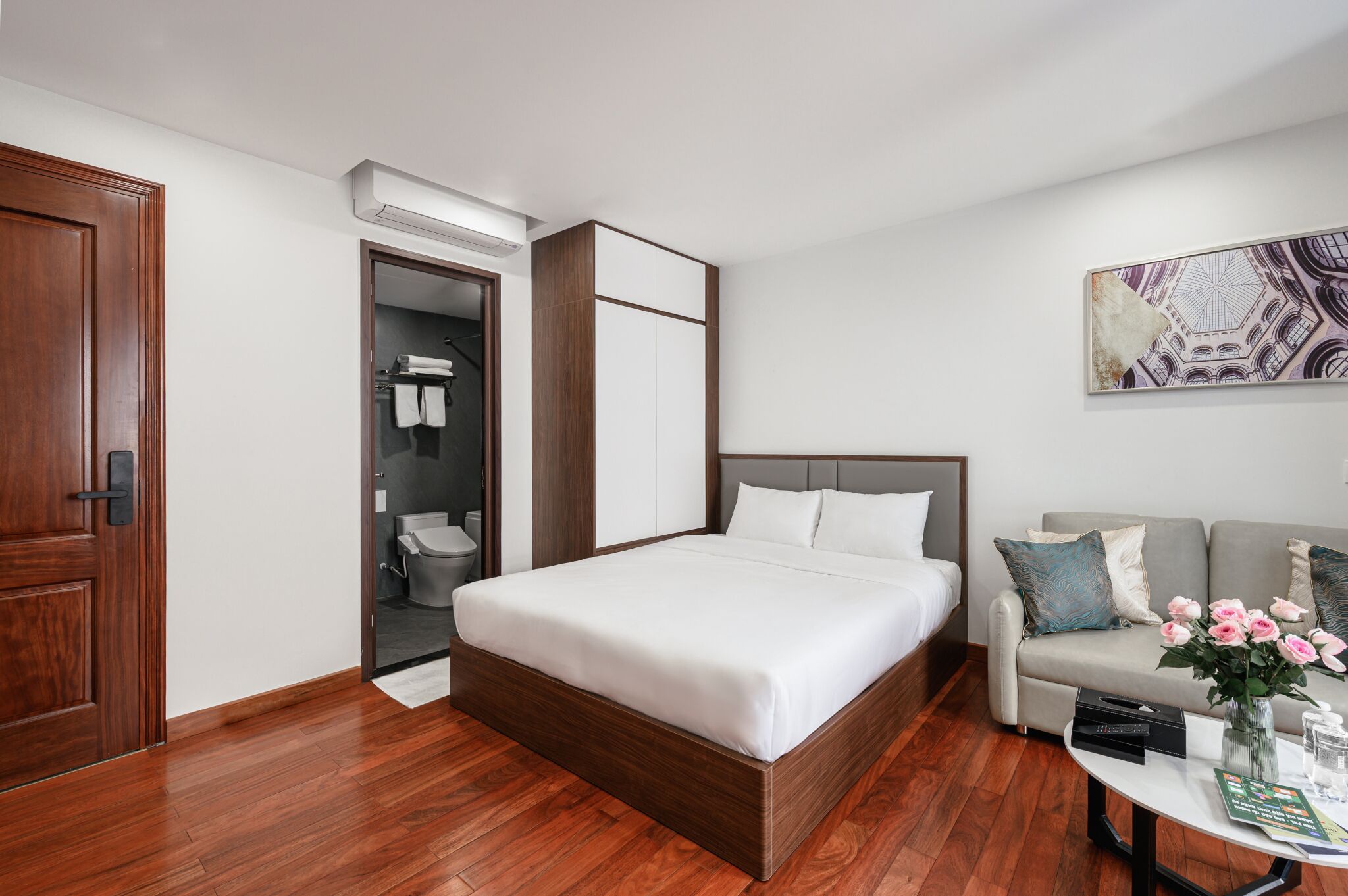 Une chambre d’hôtel avec un plancher en bois, un lit avec une literie blanche, un canapé gris agrémenté de coussins décoratifs, une table basse avec un vase de fleurs et un tableau au mur.