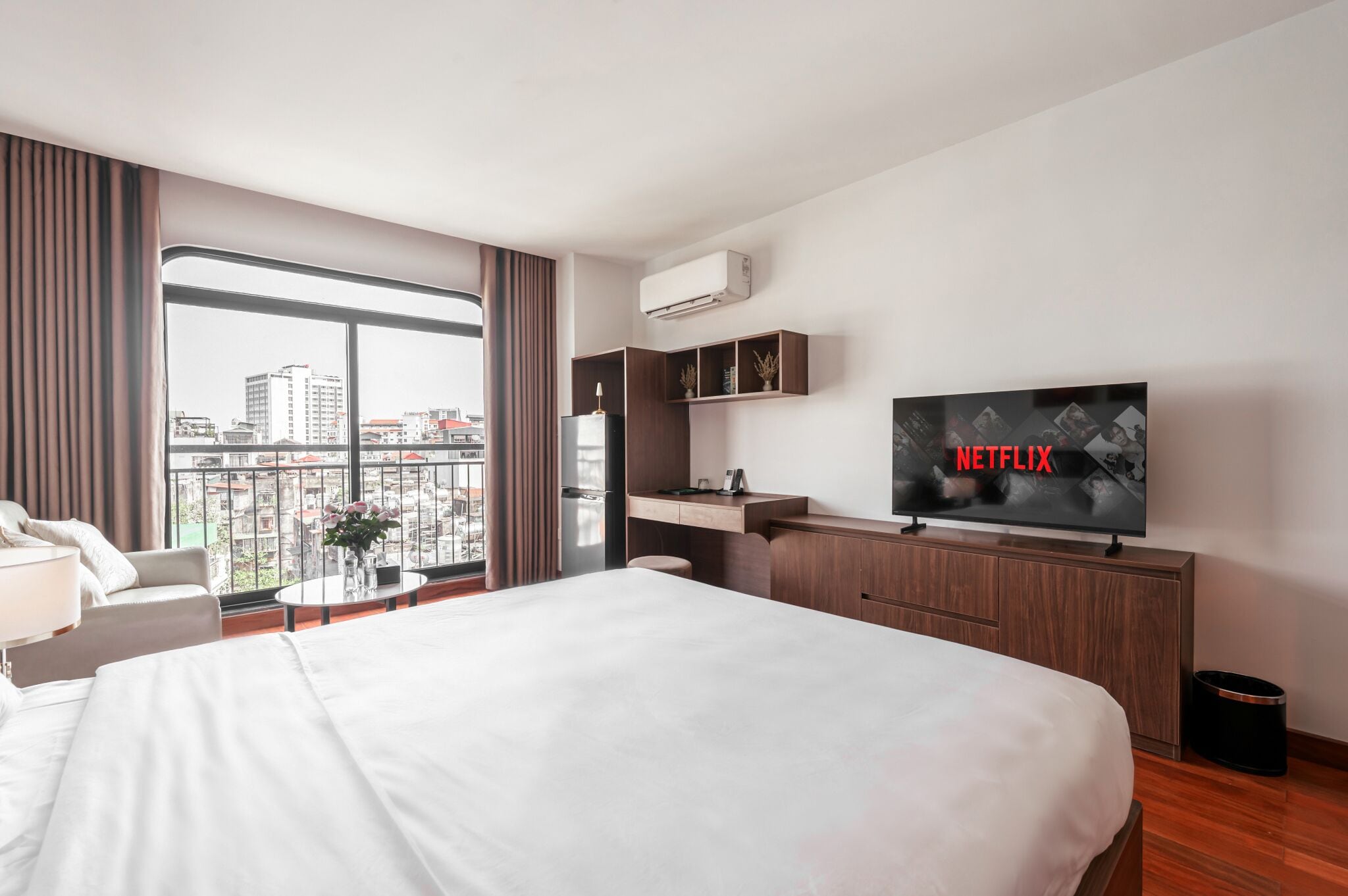 A hotel room with a large bed, a desk, a TV displaying 'NETFLIX,' and a view of the city through large windows.