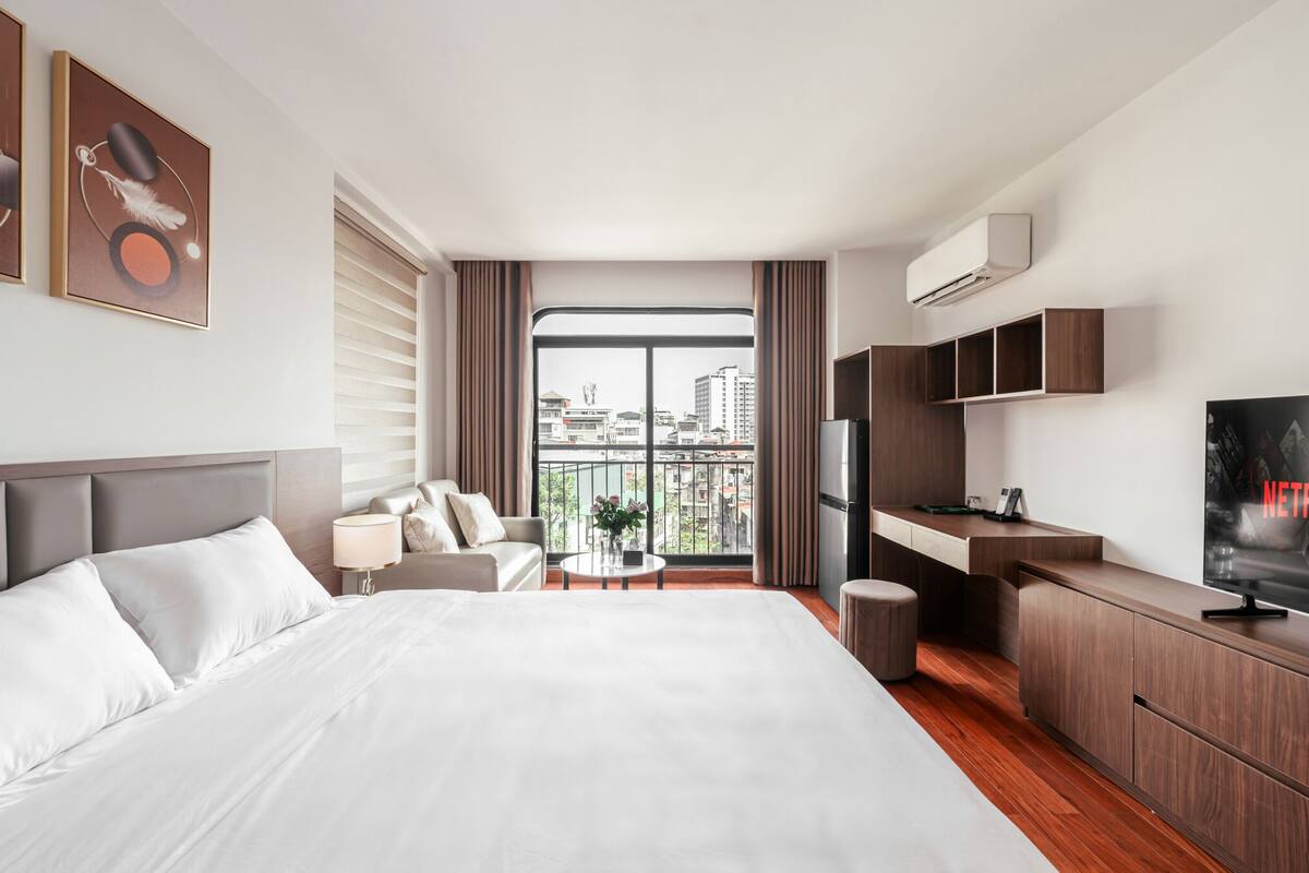 A hotel room with a large bed, a sofa, a TV, a desk, and a balcony with a city view.