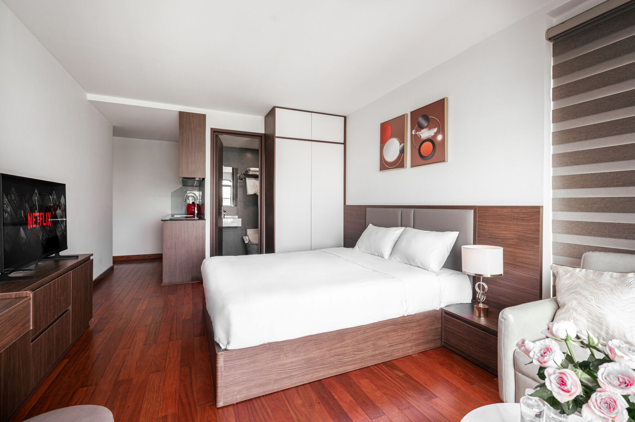 Habitación de hotel moderna con suelo de madera, cama con ropa blanca, televisor, escritorio y baño visible a través de una puerta abierta.