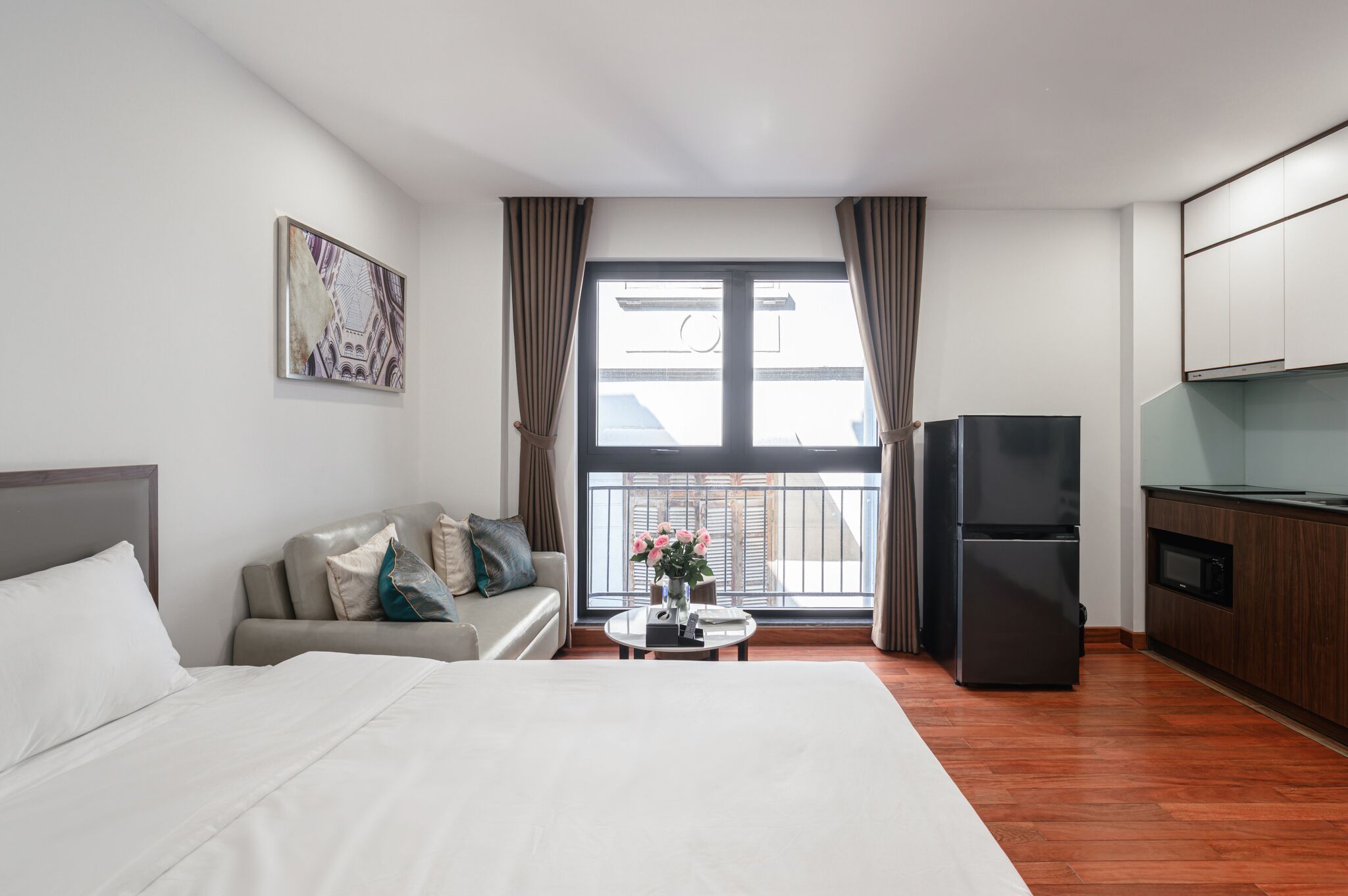 Habitación de hotel con cama, sofá, mesa de comedor con flores, refrigerador y zona de cocina.