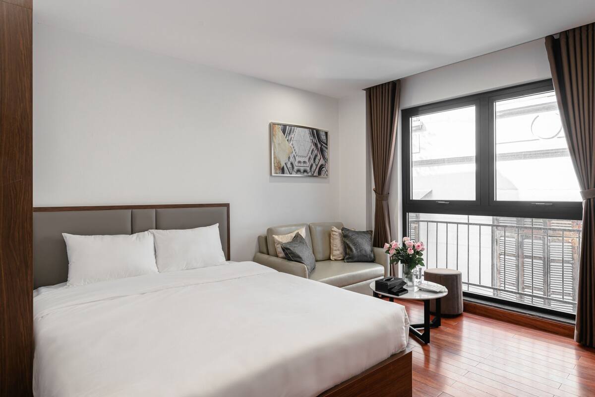 A hotel room with a bed, a sofa, a small table with a vase of flowers, and a window with curtains.