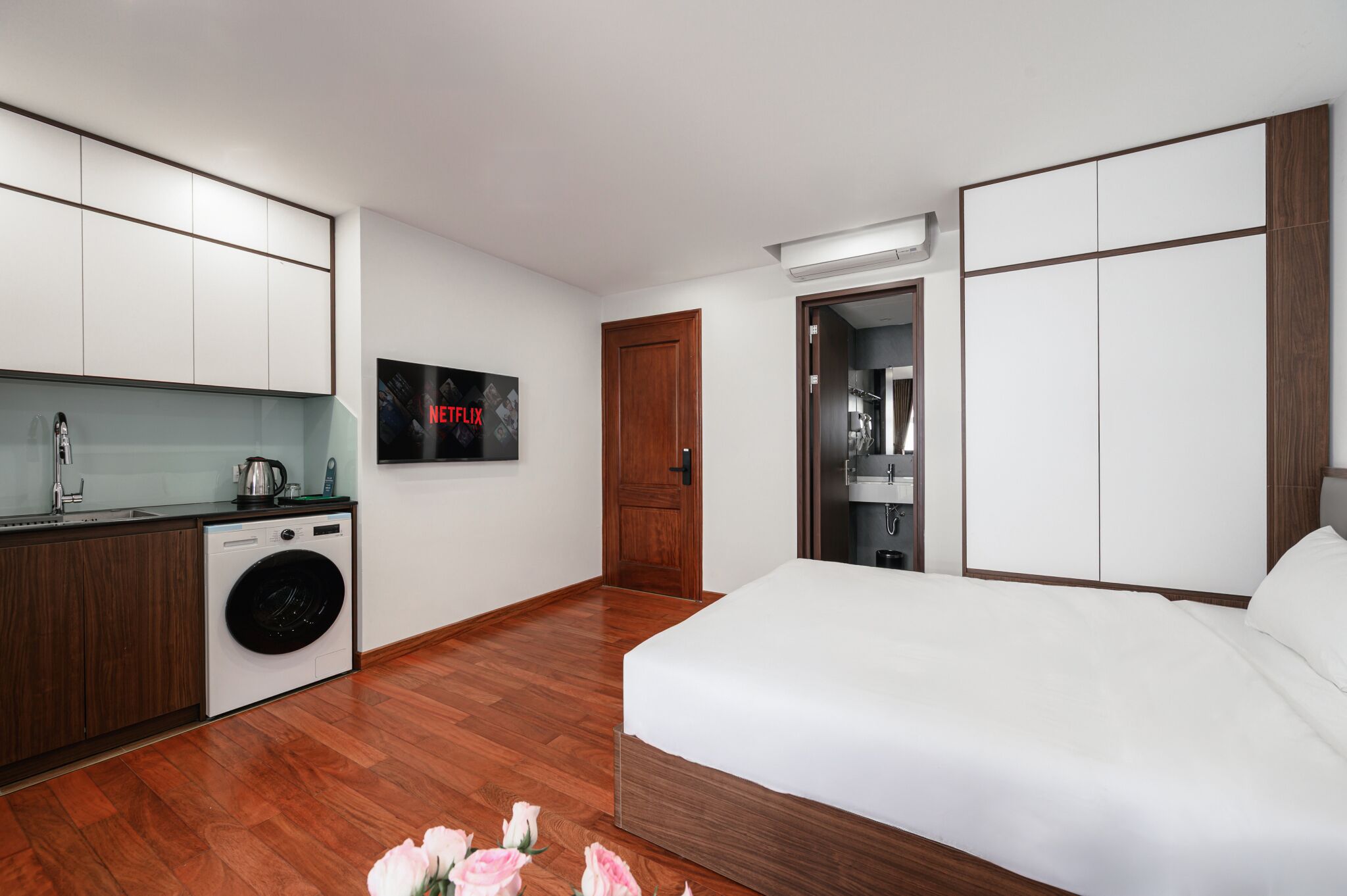 A modern hotel room with a bed, a washing machine, a kitchenette, and a door leading to a bathroom.