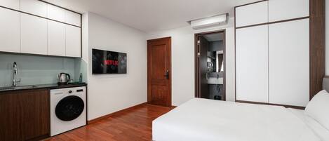 A modern hotel room with a bed, a washing machine, a kitchenette, and a door leading to a bathroom.