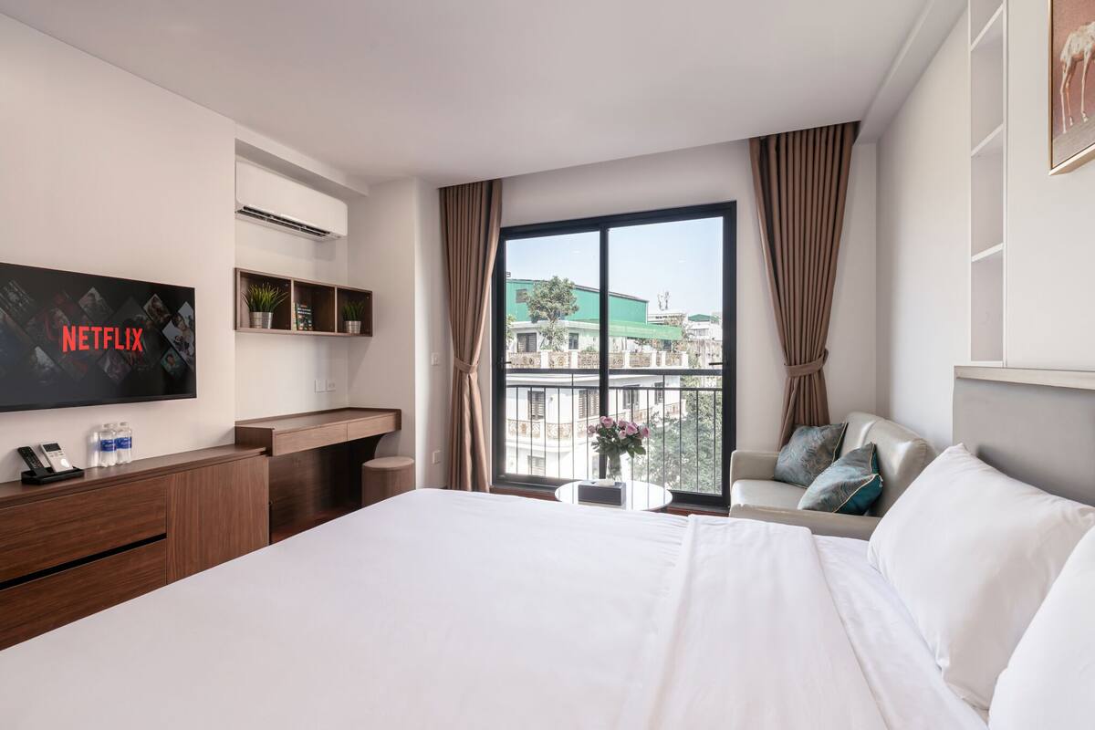 A hotel room with a large bed, a TV mounted on the wall, a desk with a phone and water bottles, a balcony with a view of buildings, and a sofa with pillows.