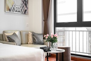 Ein modernes Hotelzimmer mit einer Couch, einem kleinen runden Tisch mit Blumenvase, einer Fernbedienung und einem Fenster mit Blick auf Gebäude.