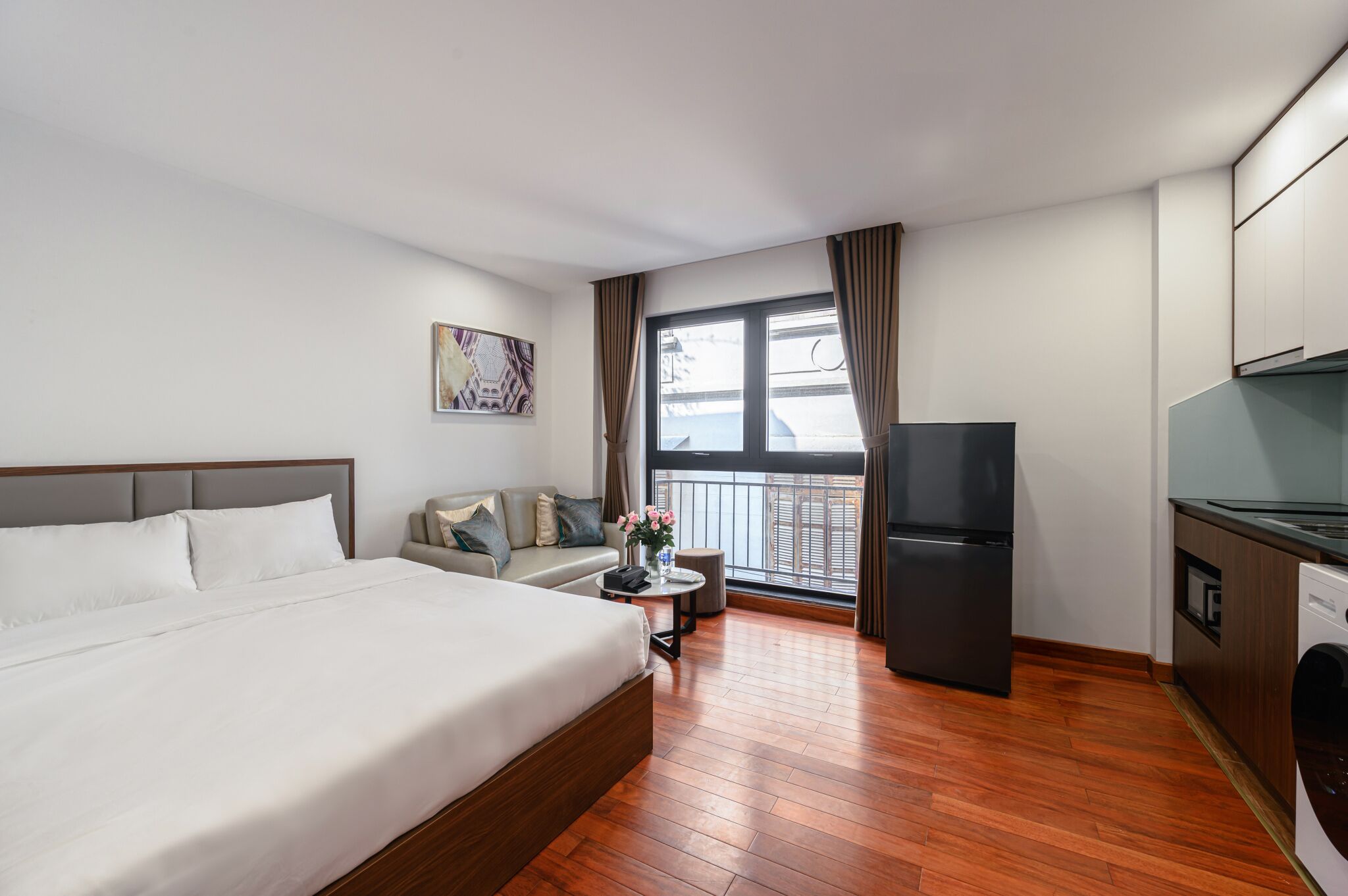 A hotel room with a bed, a sofa, a small table with a vase, a refrigerator, and a washing machine.