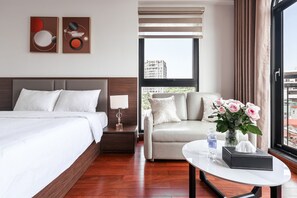 A hotel room with a bed, a sofa, a table with a vase of flowers, and a window with a view of buildings. - Luxe Paradise Suites Doi Can (Ba Dinh)
