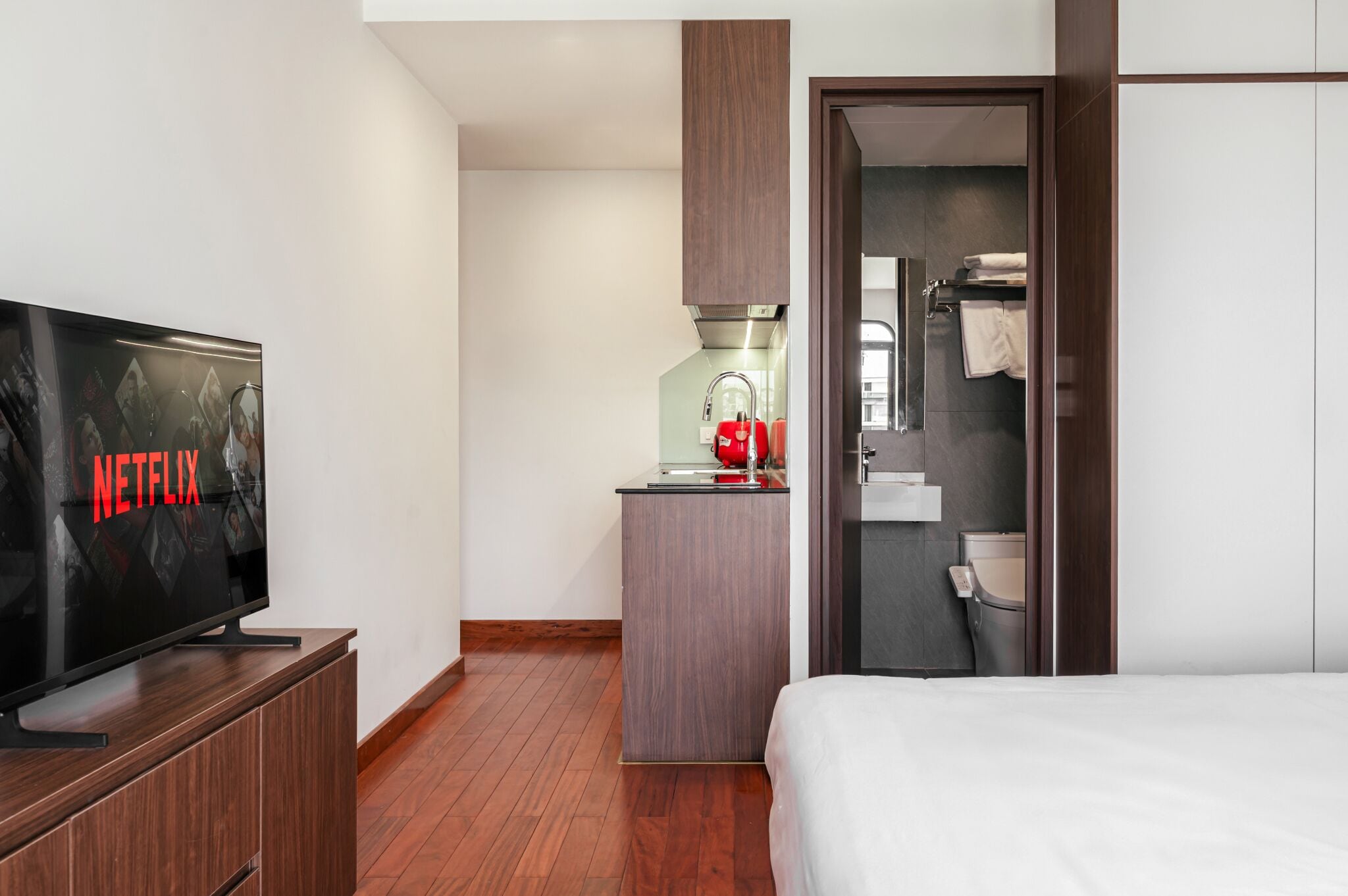 A hotel room with a bed, a TV displaying 'NETFLIX,' a kitchenette, and a bathroom.