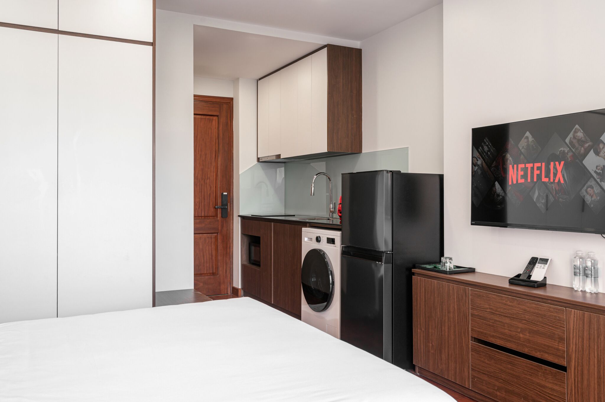 Ein modernes Hotelzimmer mit Bett, Waschmaschine, Kühlschrank, einem Holzschrank und einem an der Wand befestigten Fernseher, der das Netflix-Logo zeigt.