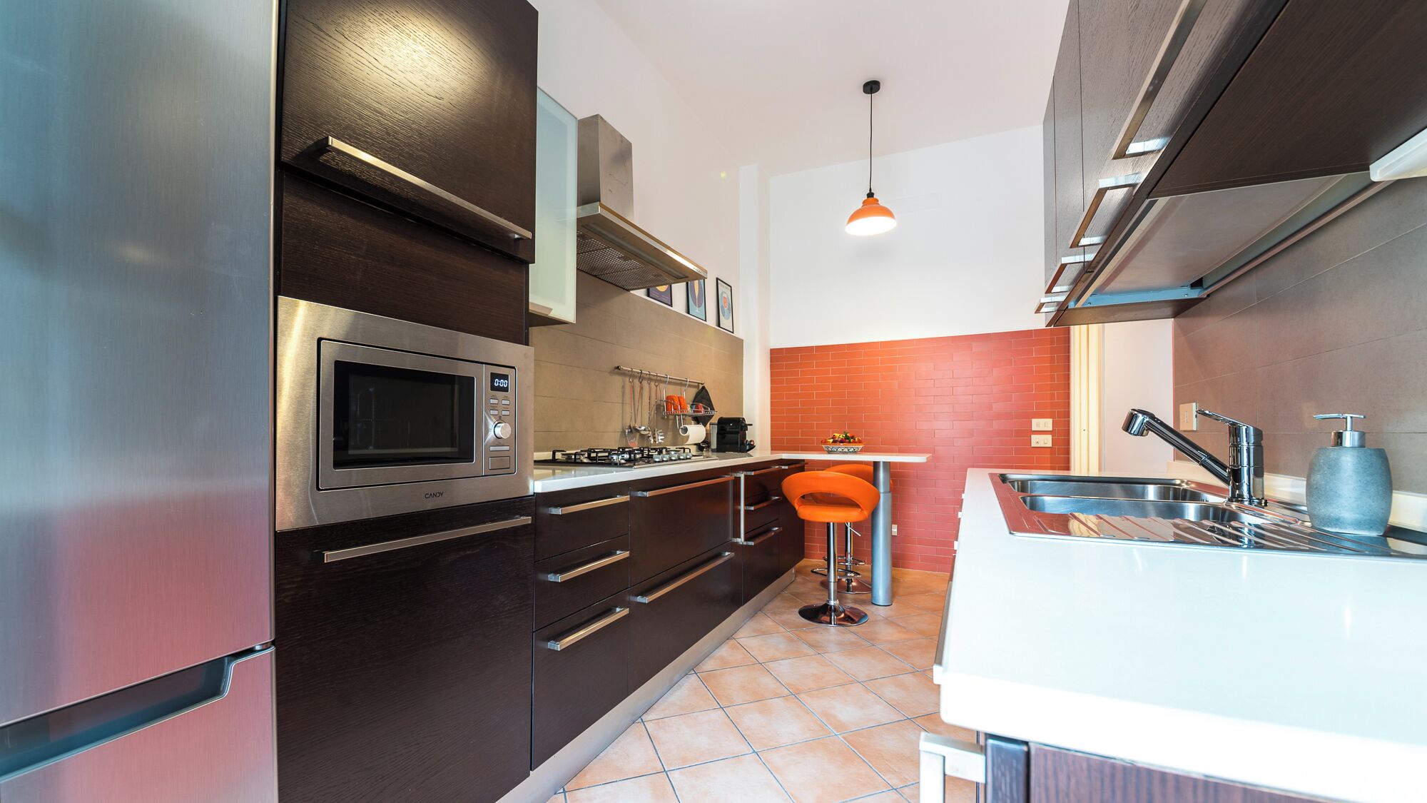 Una cocina moderna con armarios oscuros, electrodomésticos de acero inoxidable, una encimera blanca y una pared decorativa de ladrillo rojo.