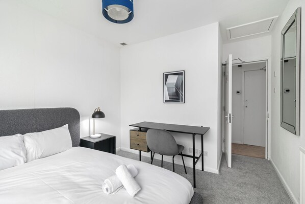 A hotel room with a bed, a desk, a chair, a lamp, and a door leading to a bathroom. - Refurbished 4BDR Home in Harborne Birmingham (Birmingham)