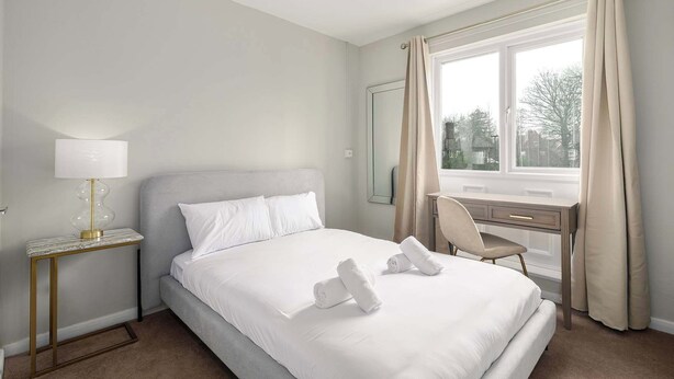 Quarto de hotel com cama, escrivaninha, cadeira, abajur e janela com cortinas.