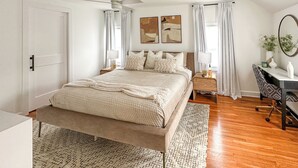 Une chambre à coucher comprenant un lit, des tables de chevet, un bureau, une chaise, un ventilateur de plafond et des œuvres d’art accrochées aux murs.