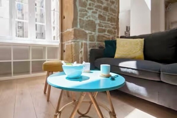 A room with a stone wall, a grey sofa with a yellow pillow, a blue table with a white bowl and a cup, and a wooden floor. - Plein center, Place des Jacobins, Lyon (Lyon)