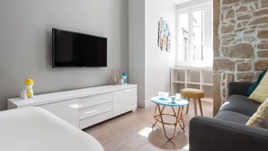 A modern living room with a flat-screen TV, white furniture, a stone accent wall, and a view through an open door.