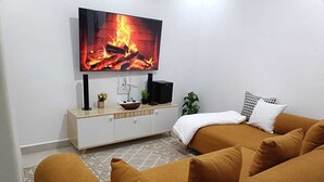Un salon moderne comprenant un canapé, un téléviseur à écran plat diffusant une image de cheminée, un petit meuble et une plante.