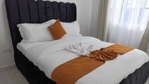 Un lit bien fait, avec une literie blanche et marron, une tête de lit sombre et une fenêtre ornée de rideaux légers.