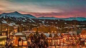 A town with snow-covered mountains in the background, buildings with snow on their roofs, and lights on in the foreground. - Lux Slope-side 1BR.2BAw/FullKitchen, 1 King, 1 Queen Sofa  @ Grand Lodge Peak 7 (Breckenridge)