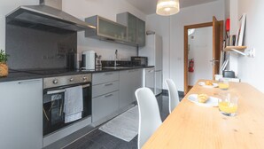 Et moderne køkken med indbygget ovn, mikroovn og emhænde i rustfrit stål. Der er et spisebord med stole og to glas appelsinjuice.