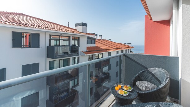 En balkon med fletmøbler, gelænder af glas og et bord med mad og drikke.