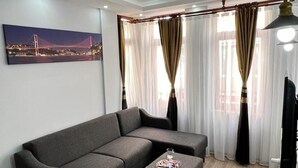 En moderne stue med en grå sofa og skammel, et indrammet bybillede og lette gardiner med en bøjet gardinstang.