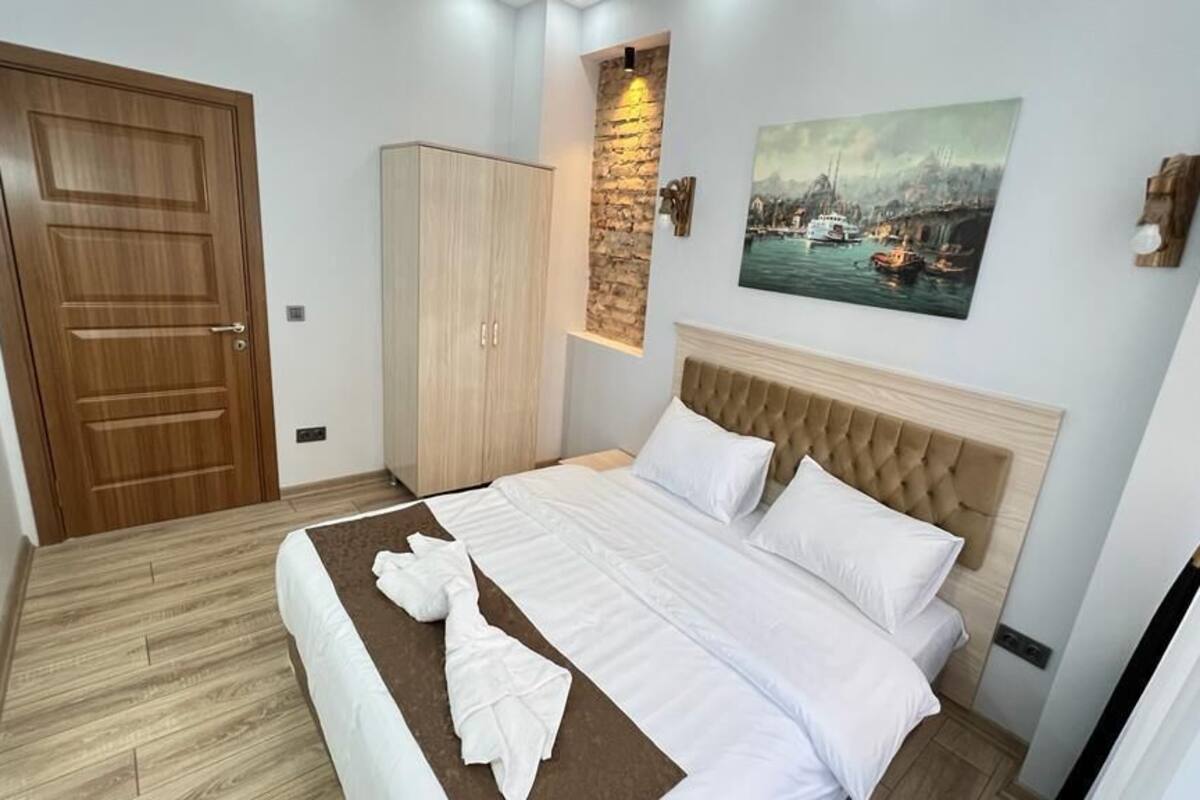 A neatly made bed with white linens, a wooden wardrobe, a painting of a harbor scene, and a stone accent wall.