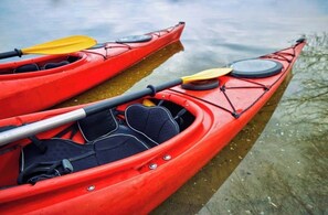 Zwei rote Kajaks mit Paddeln und Schwimmwesten liegen an einem ruhigen Gewässer festgemacht.