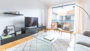 Ein modernes Wohnzimmer mit Flachbildfernseher, einem Glastisch, einer Couch und einem großen Fenster mit Vorhängen.